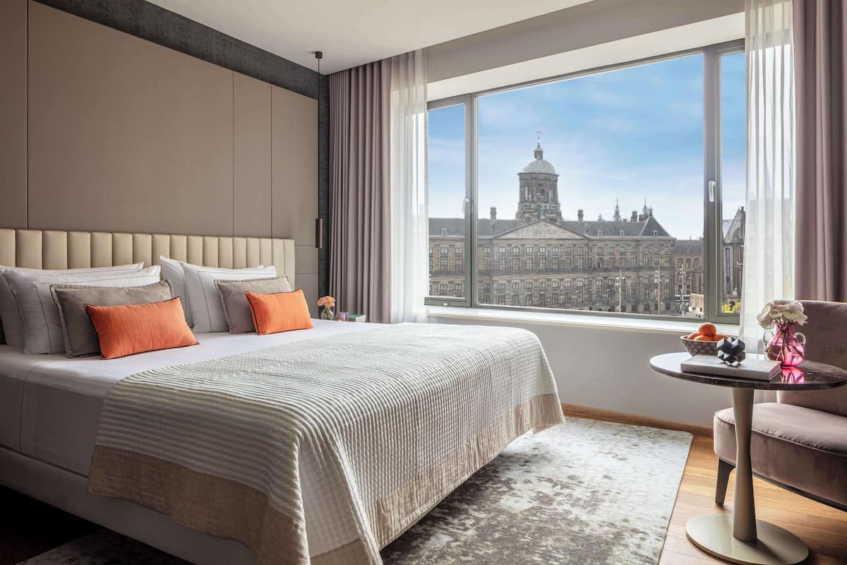 A modern hotel room with a large bed, a small round table, and a view of a historic building through a large window.