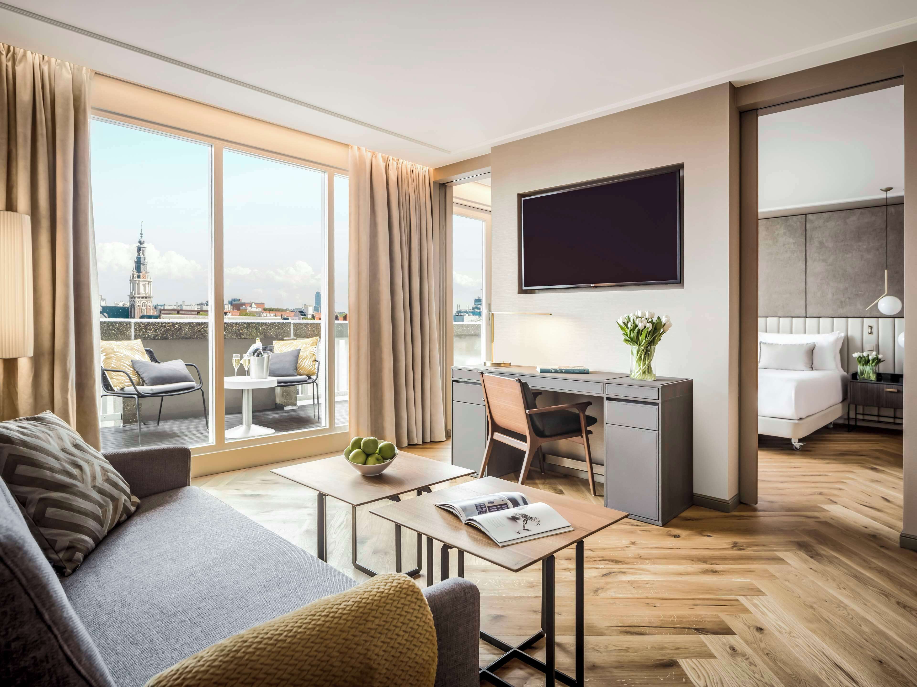 Een moderne hotelkamer met een bank, een eettafel, een bureau en een televisie. De kamer heeft grote ramen met gordijnen, uitzicht op de stad en een houten vloer.