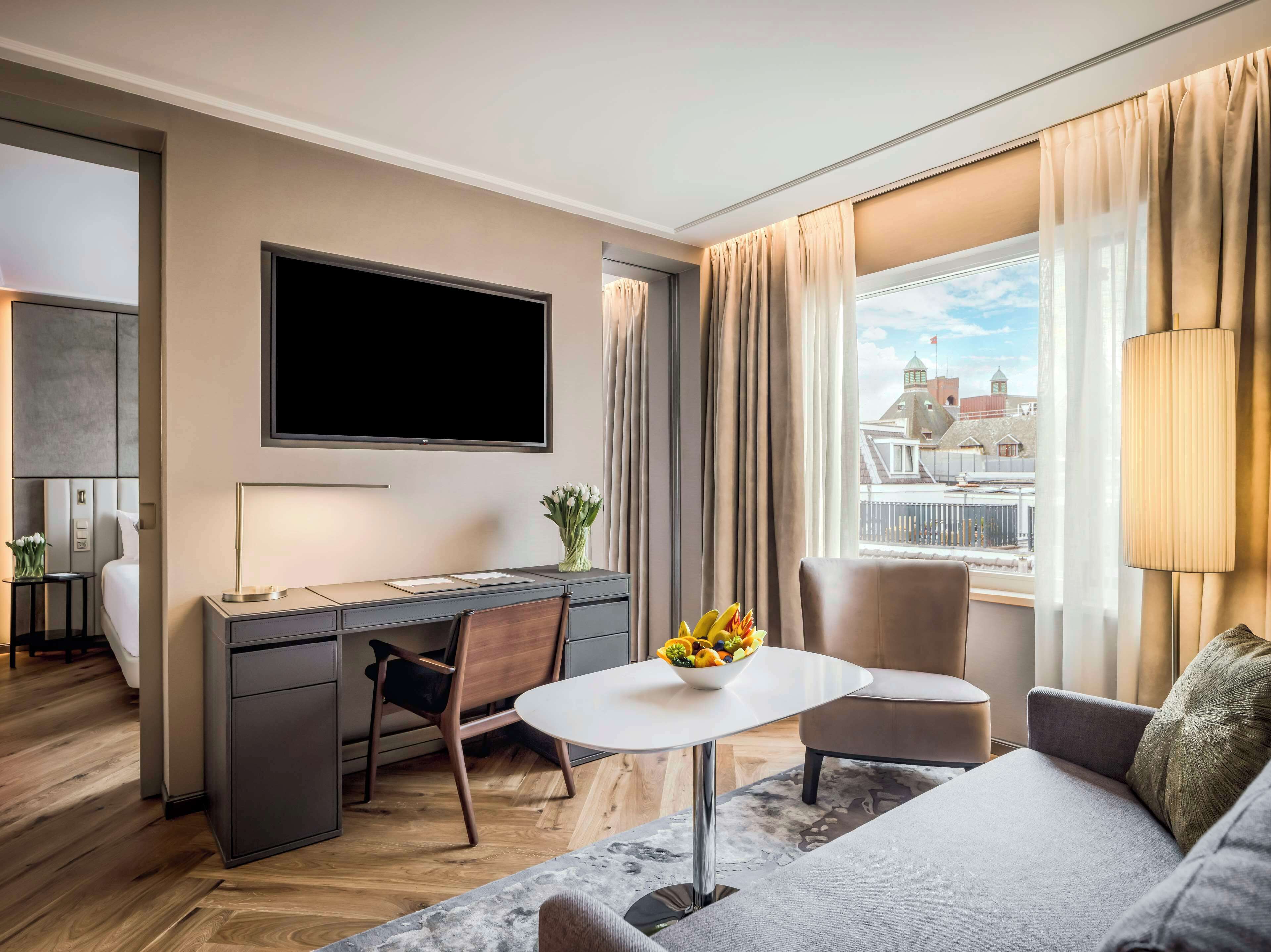 Ein modernes Hotelzimmer mit Flachbildfernseher, Schreibtisch mit Stuhl, einer Couch und einem Esstisch mit Obstschale.