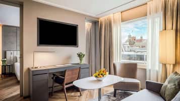 Ein modernes Hotelzimmer mit Flachbildfernseher, Schreibtisch mit Stuhl, einer Couch und einem Esstisch mit Obstschale.