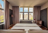 A hotel room with a large bed, a chair, a small table with a vase, and a view of city buildings through the window.