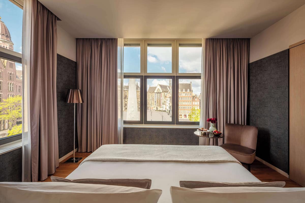 A hotel room with a large bed, a chair, a small table with a vase, and a view of city buildings through the window.