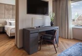 A modern hotel room with a large bed, a desk with a chair, a flat-screen TV, and a view of a cityscape through a window.