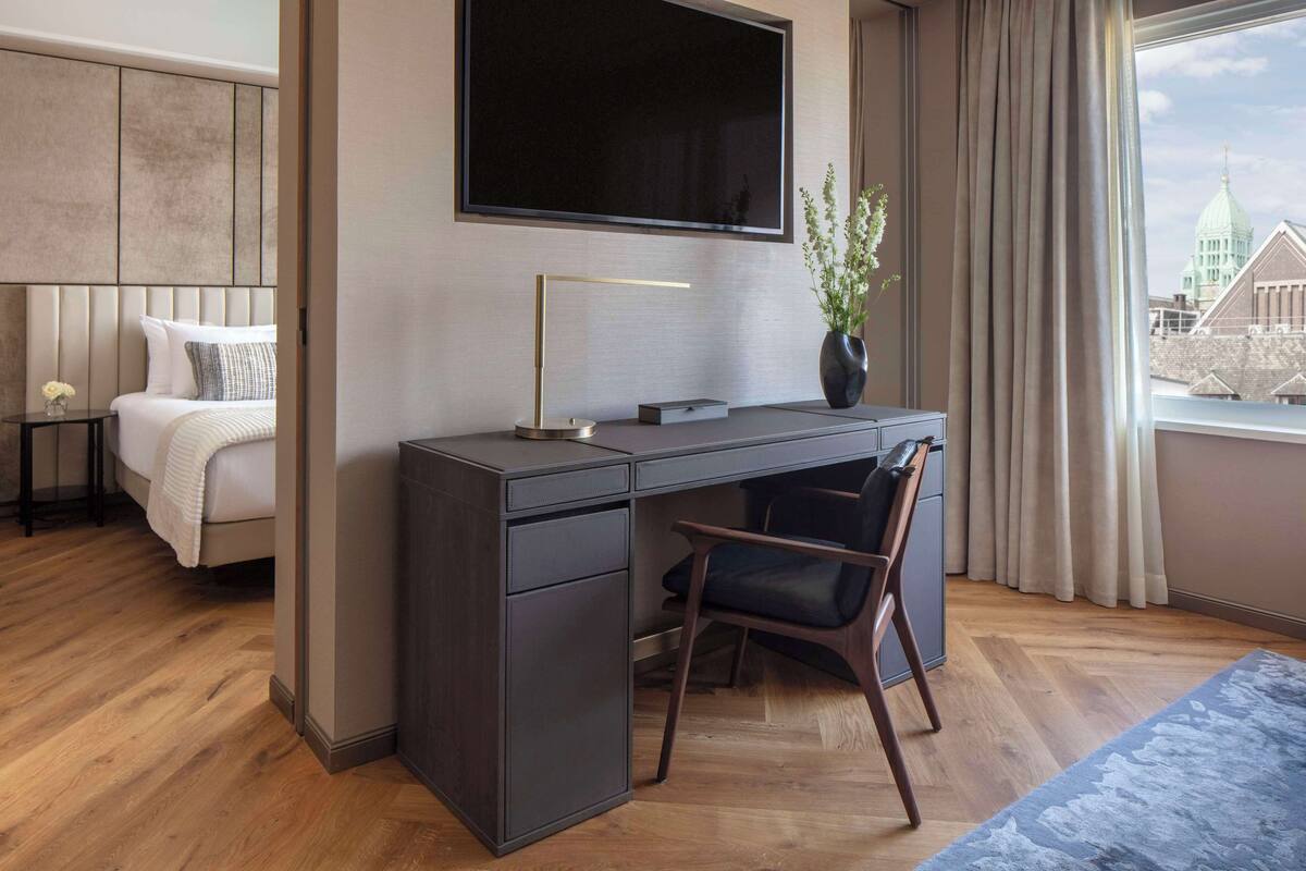 A modern hotel room with a large bed, a desk with a chair, a flat-screen TV, and a view of a cityscape through a window.