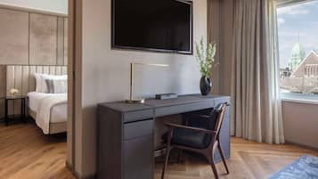 A modern hotel room with a large bed, a desk with a chair, a flat-screen TV, and a view of a cityscape through a window.