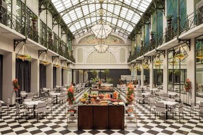 Ein geräumiger Innenhof mit Schachbrettmuster-Boden, ein zentral platziertes Buffet mit verschiedenen Speisen und eine hohe Decke mit einer Glaskuppel.