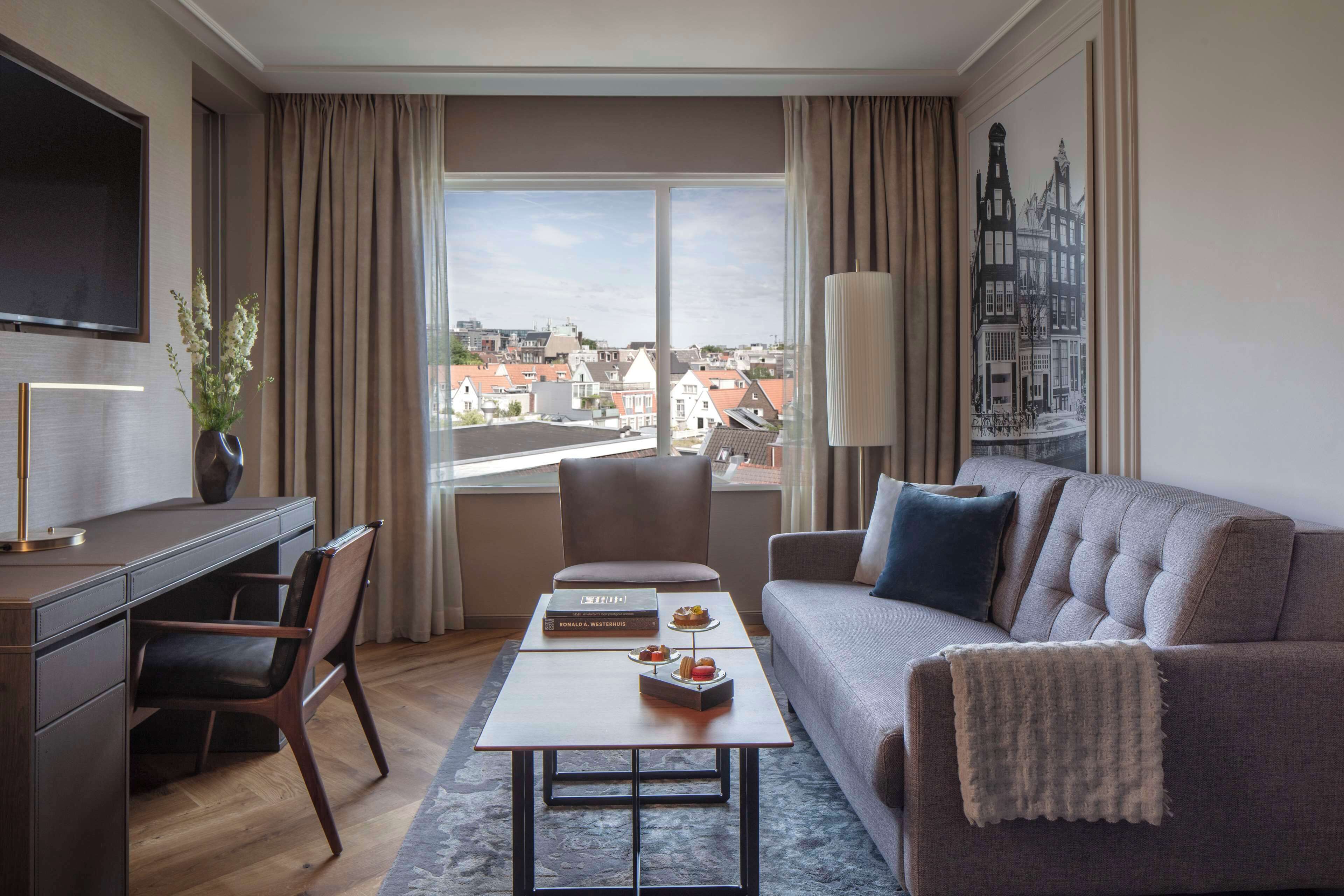 A modern hotel room with a large window, a sofa, a desk, and a coffee table.