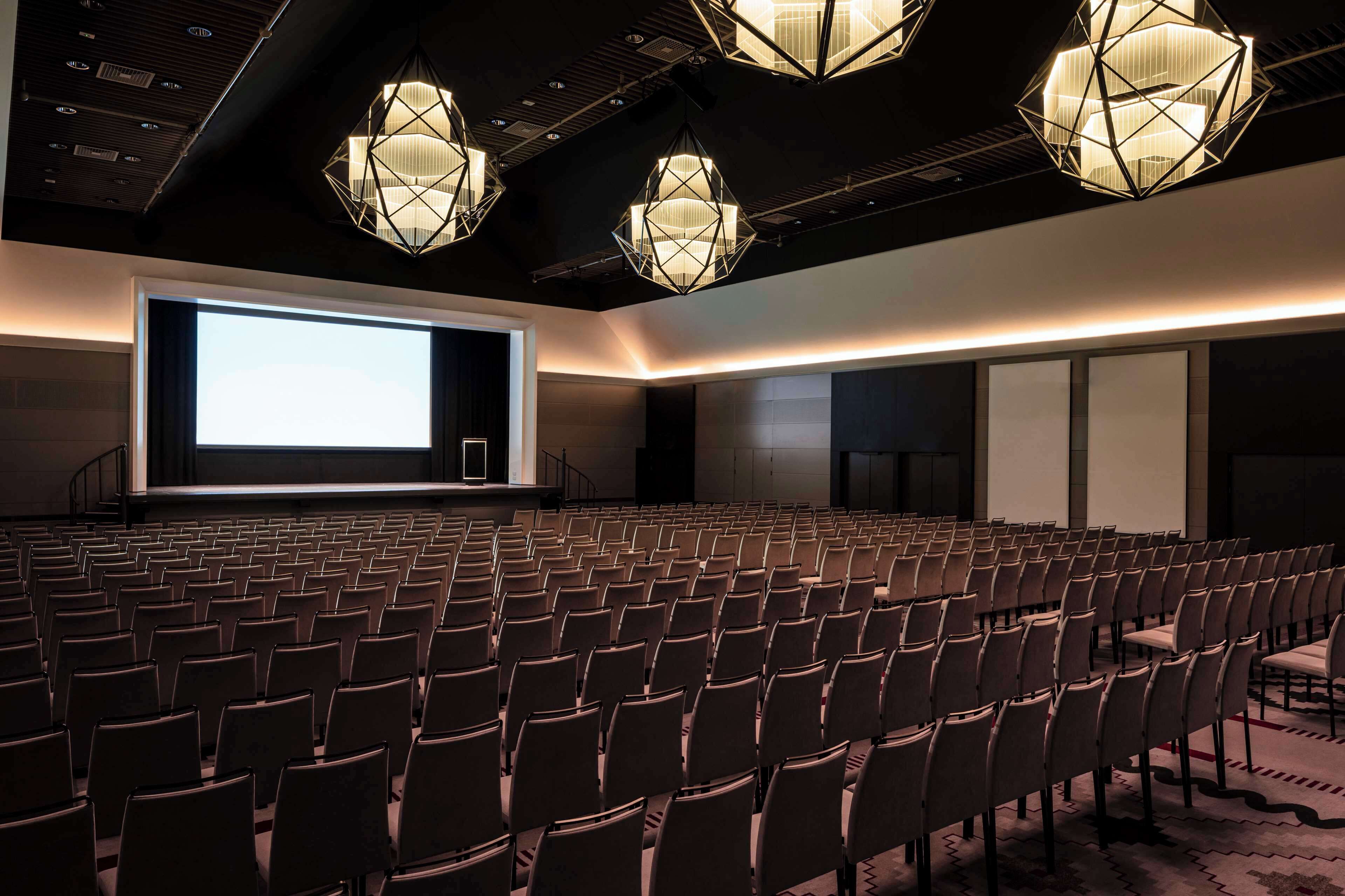 A conference room with rows of chairs, a large screen, and geometric pendant lighting.