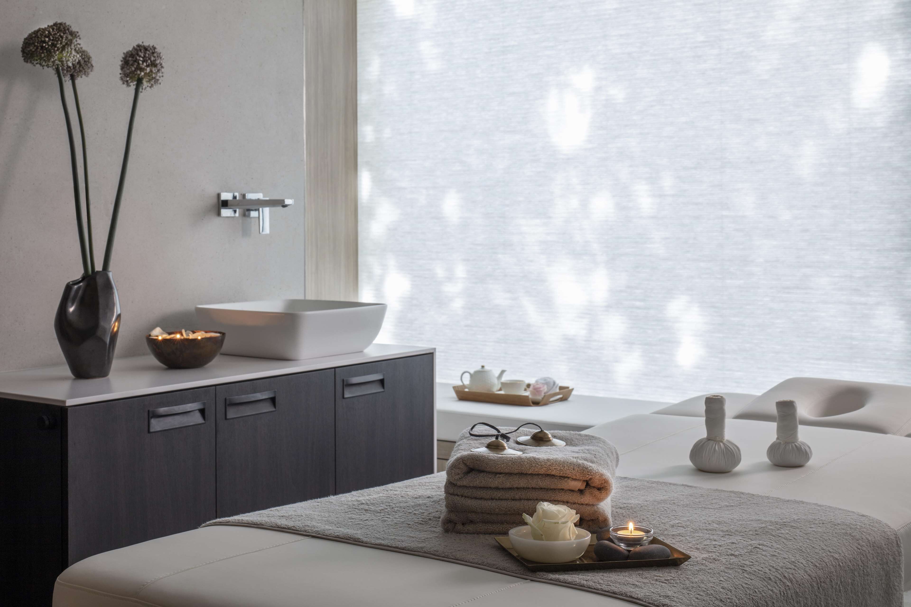 A modern spa room with a large window, a sink, a cabinet, towels, a bowl, and a tray with a candle.