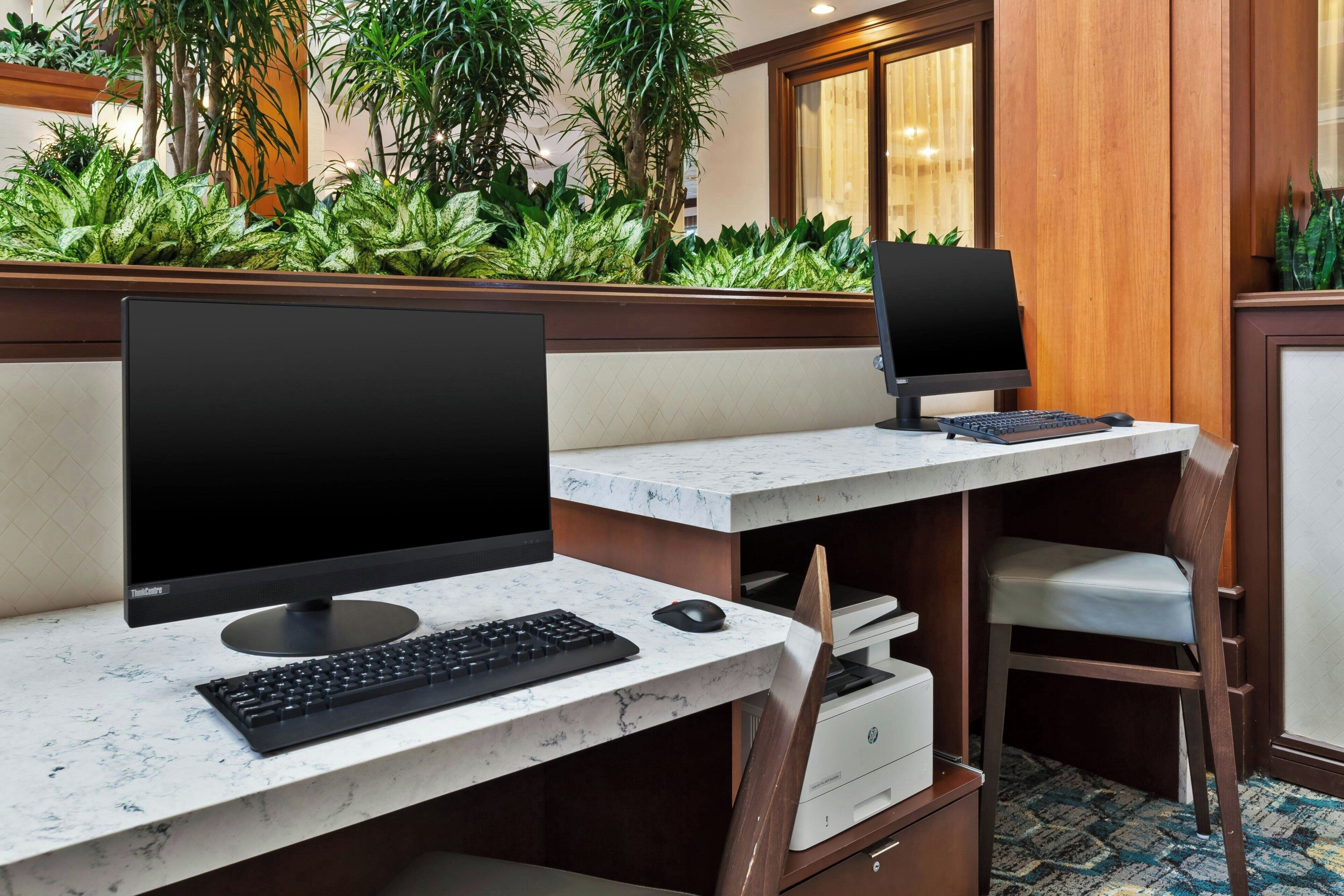Un espacio de trabajo en un hotel con dos escritorios de ordenador, cada uno con monitor, teclado y ratón. Se ven plantas al fondo, y la habitación tiene un diseño de paneles de madera.