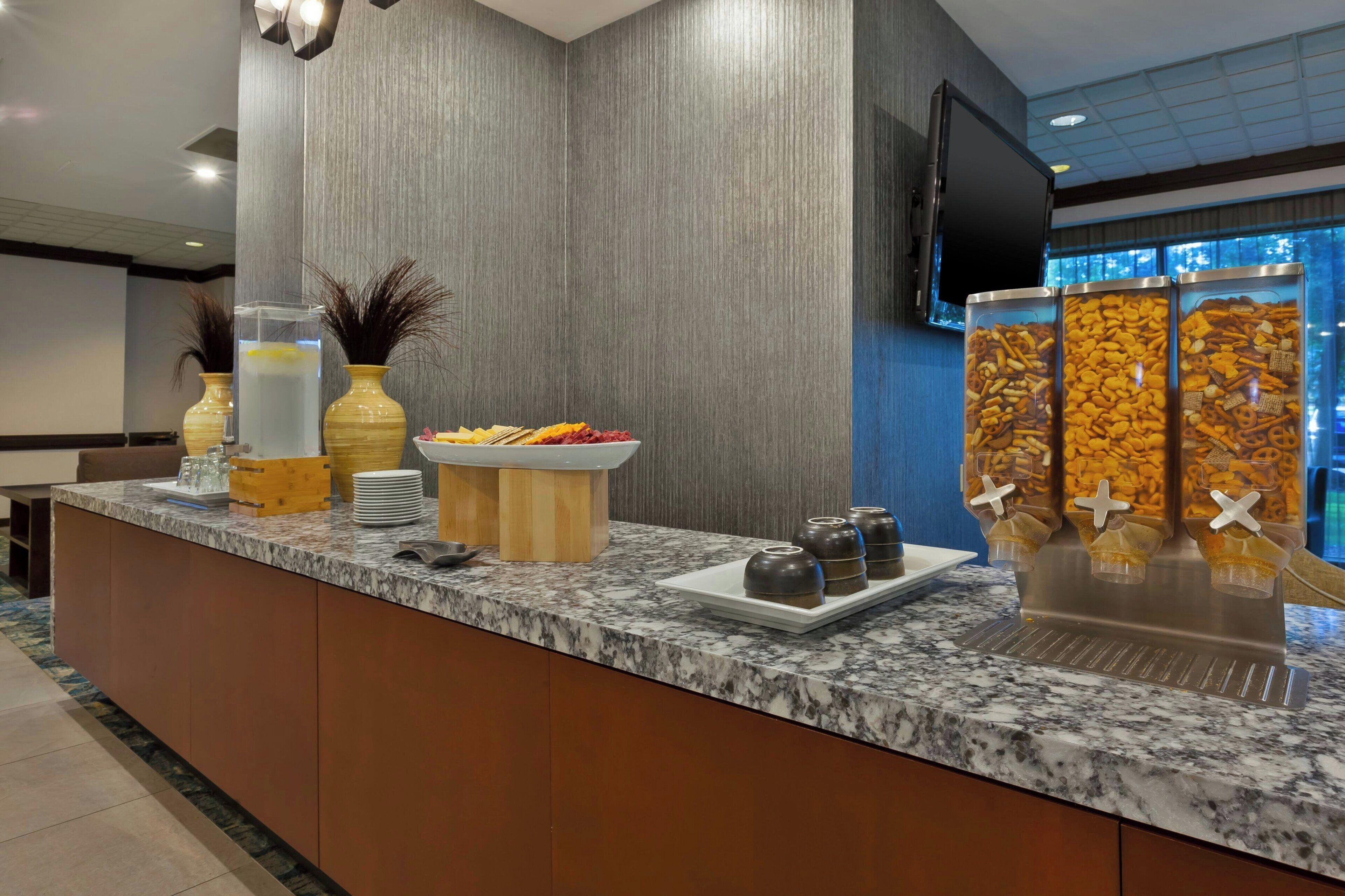 Uma área de recepção de hotel com balcão de mármore, uma cesta de frutas, dispensador de água e máquinas de lanches.