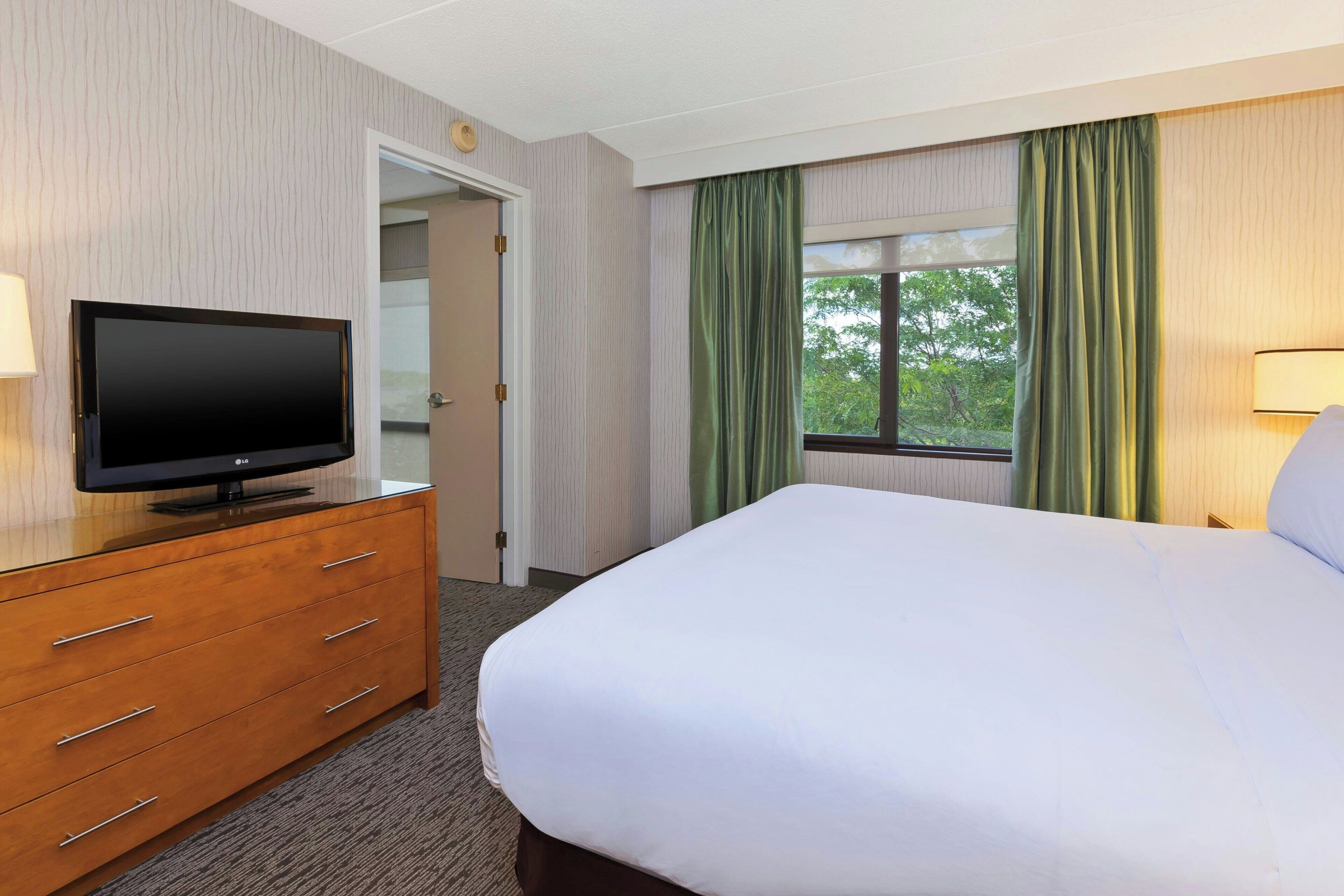 A hotel room with a bed, a television on a wooden dresser, a window with curtains, and a door.