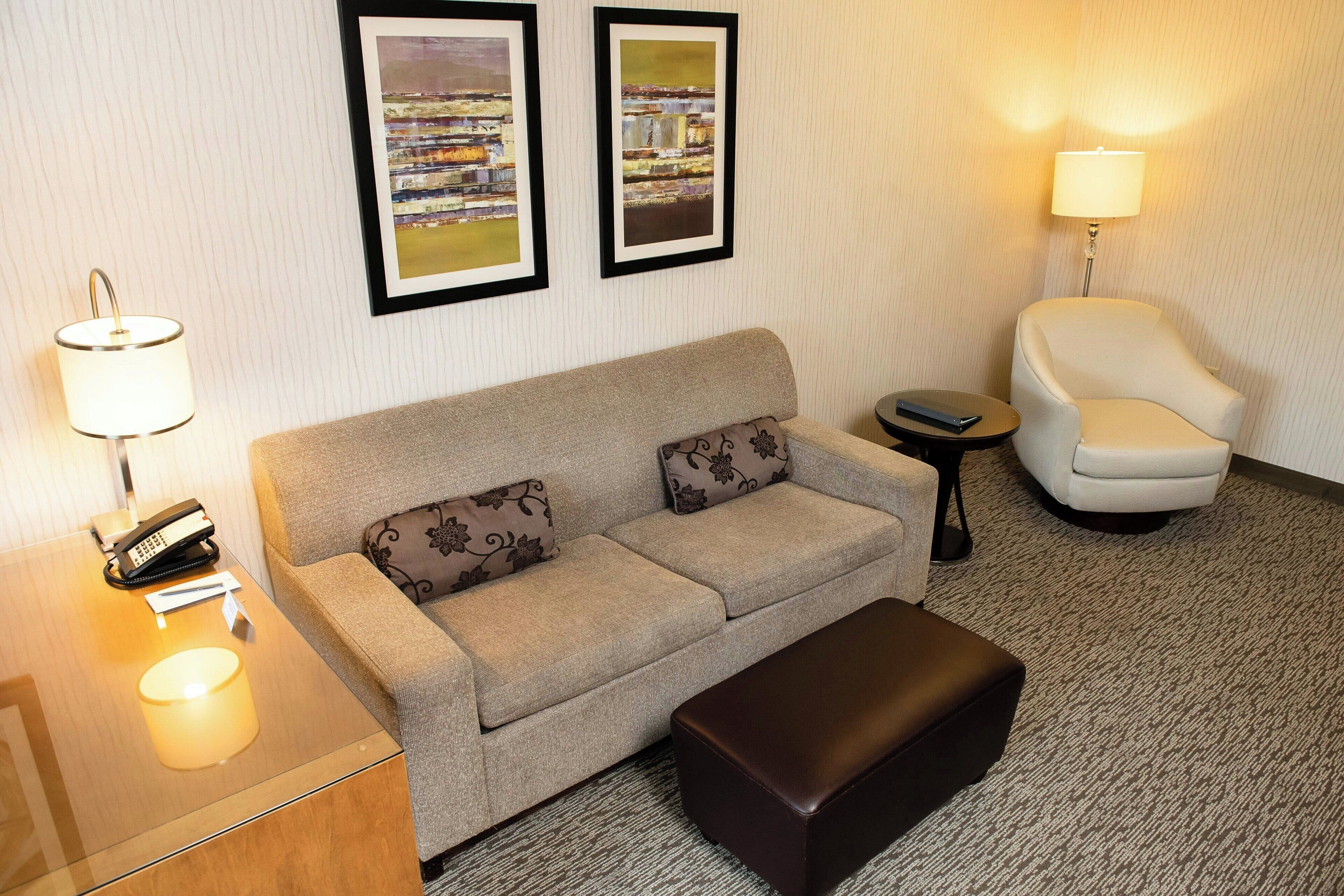 A hotel room with a sofa, a chair, a small table with a lamp, and two framed pictures on the wall.