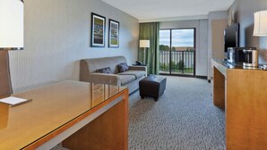 A hotel room with a sofa, ottoman, desk, and a TV. - Hotel Auburn Hills (Auburn Hills)