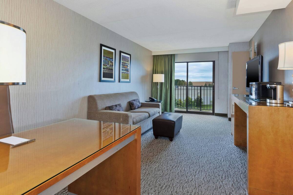 A hotel room with a sofa, ottoman, desk, and a TV.