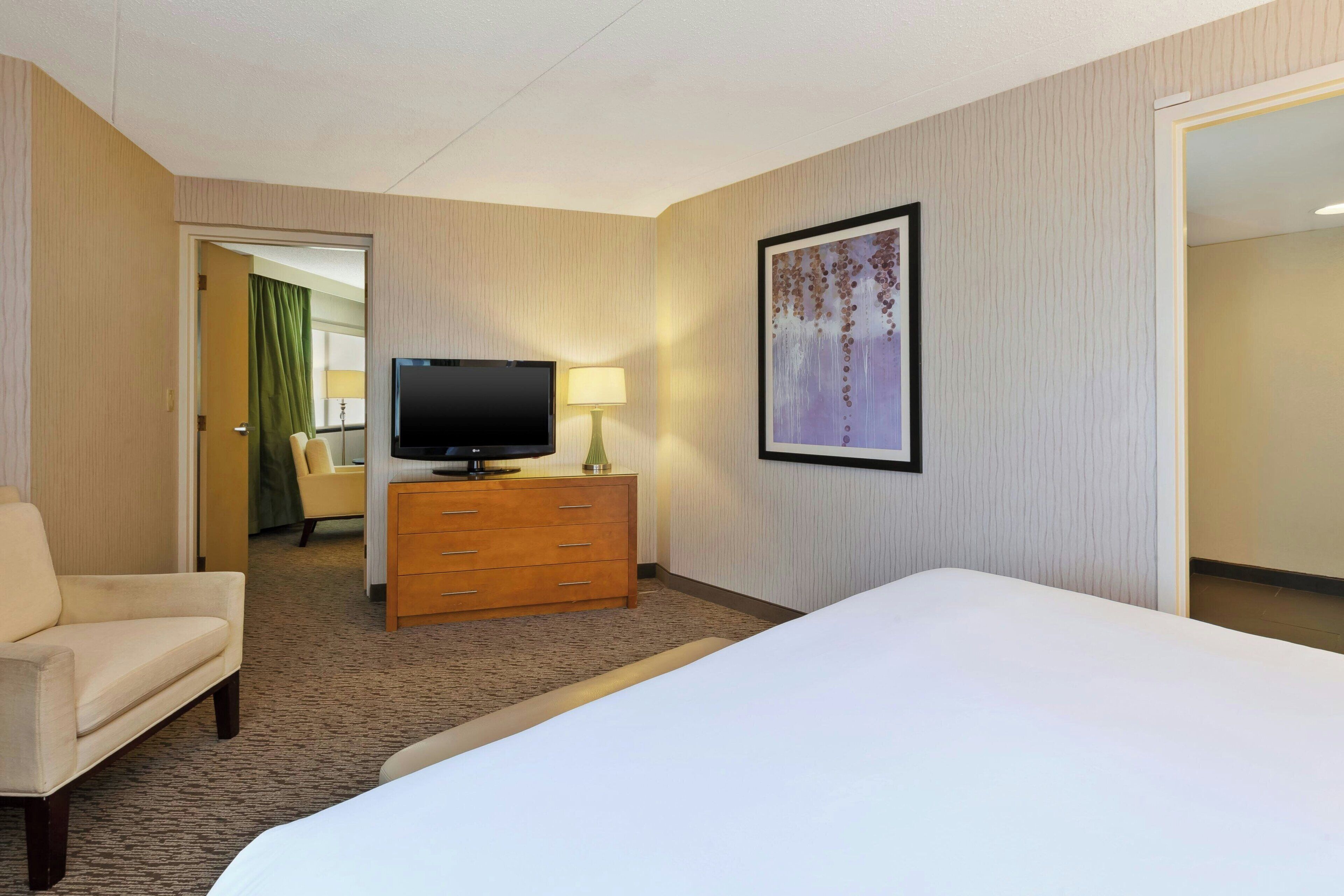 A hotel room with a bed, a television on a wooden dresser, a chair, and a painting on the wall.