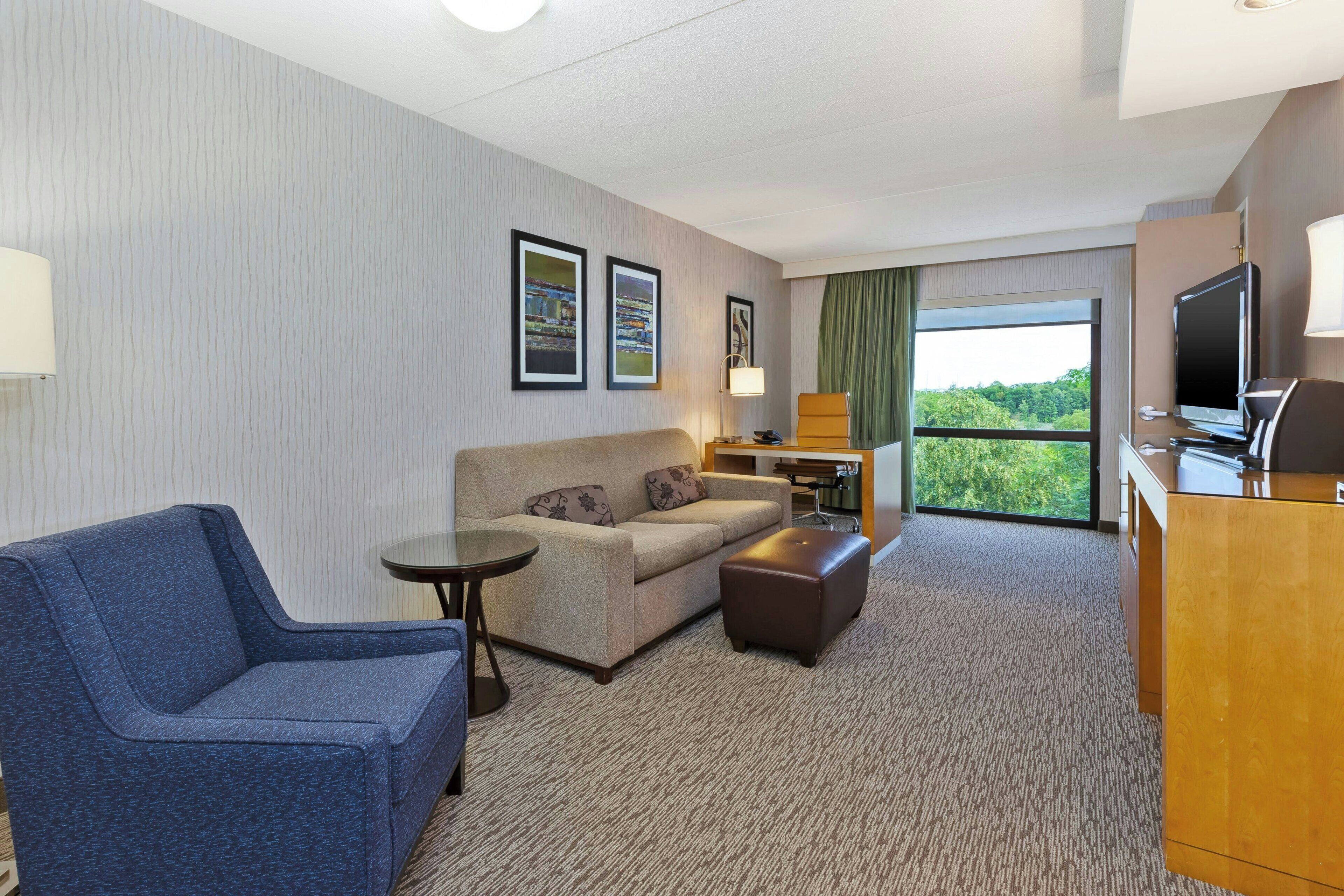 A hotel room with a sofa, a small table, a TV, and a window with a view of greenery.
