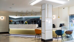 A modern reception desk with a marble top, two orange chairs, and a glass display case. - Hotel Gran Versalles (Madrid)