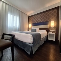 Hotellihuone, jossa on suuri sänky, yöpöytä lampun kanssa, tuoli ja ikkunasta avautuva näkymä ulos.