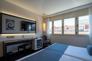 A hotel room with a bed, desk, chair, TV, and a window with a view of buildings.