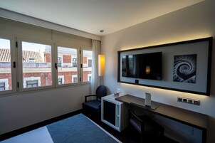 Habitación de hotel con un ventanal grande, un televisor de pantalla plana, un escritorio con lámpara y un mueble pequeño.