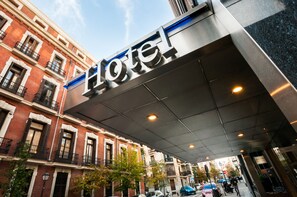 A hotel entrance with a modern sign, traditional brick buildings, and a covered walkway. - Hotel Gran Versalles (Madrid)