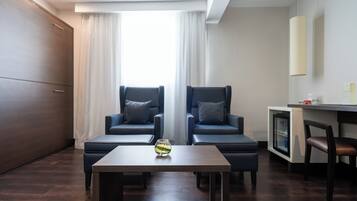 Una habitación de hotel moderna con dos sillones azul oscuro, una mesita de madera con un florero de vidrio y un minibar.