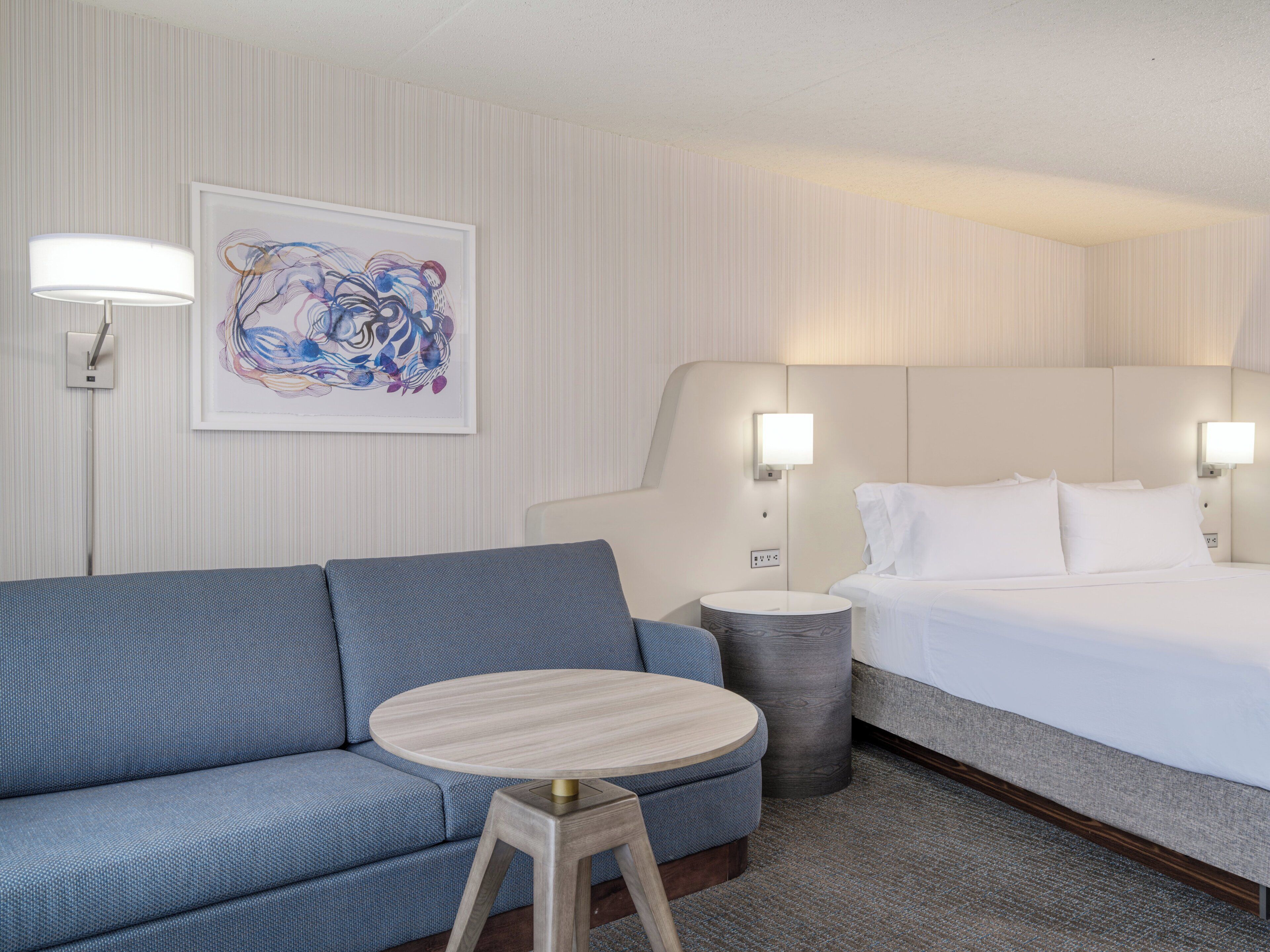Ein Hotelzimmer mit einem blauen Sofa, einem runden Holztisch, einem Bett mit weißer Bettwäsche und einem gerahmten abstrakten Gemälde an der Wand.