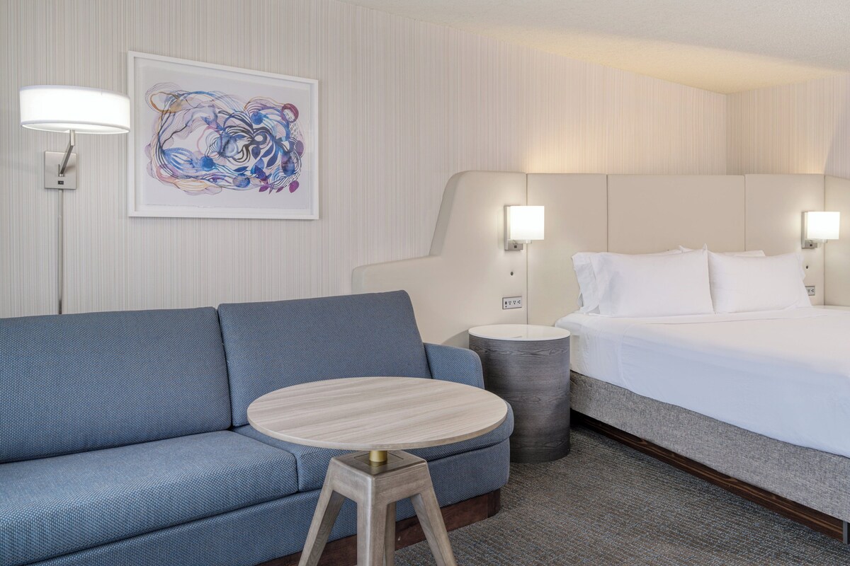 Une chambre d’hôtel avec un canapé bleu, une table ronde en bois, un lit avec du linge de maison blanc et un tableau abstrait encadré accroché au mur.