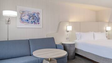 Ein Hotelzimmer mit einem blauen Sofa, einem runden Holztisch, einem Bett mit weißer Bettwäsche und einem gerahmten abstrakten Gemälde an der Wand.