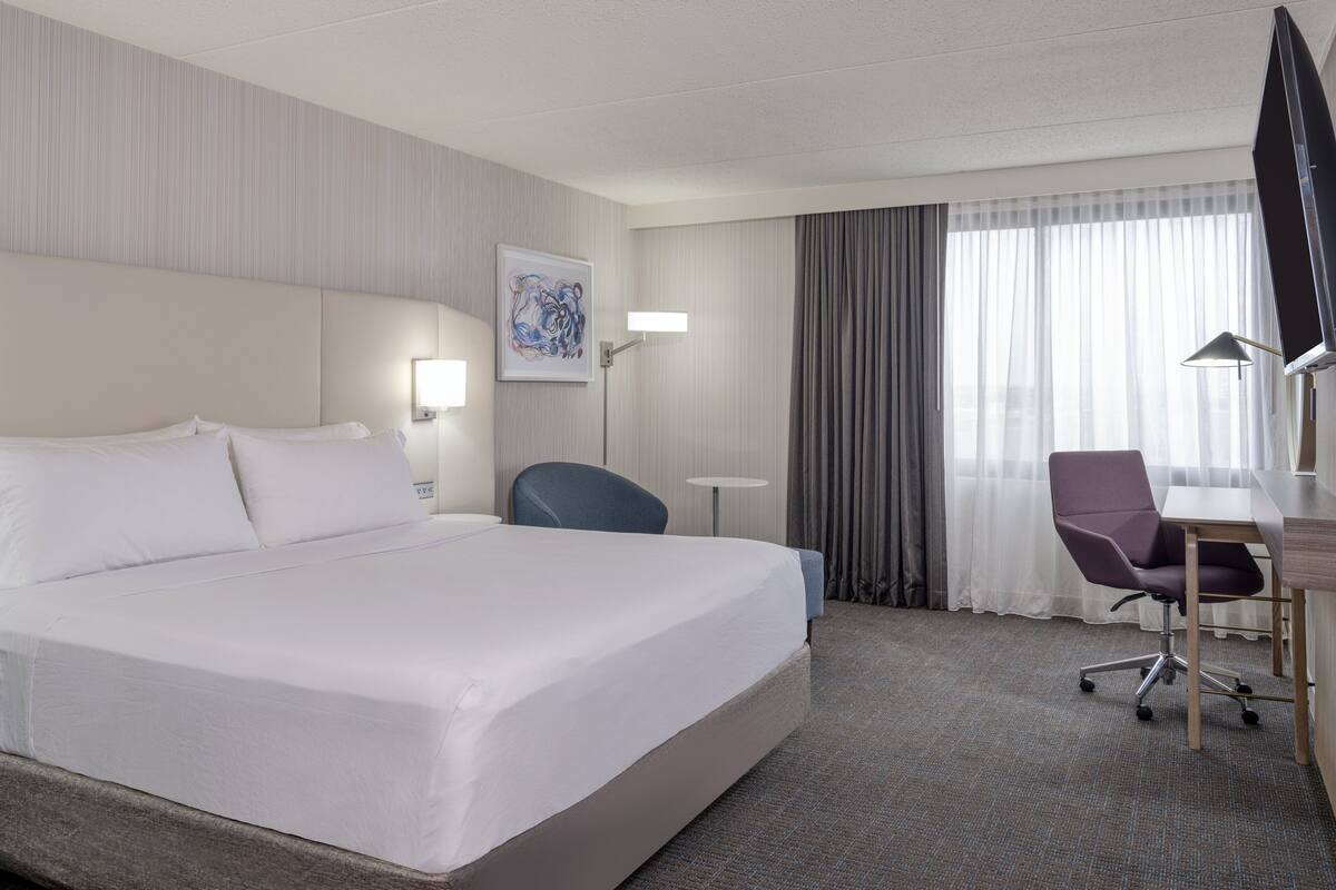 Ein Hotelzimmer mit einem großen Bett, einem Schreibtisch mit Stuhl, einem Flachbildfernseher und einem Fenster mit Vorhängen.