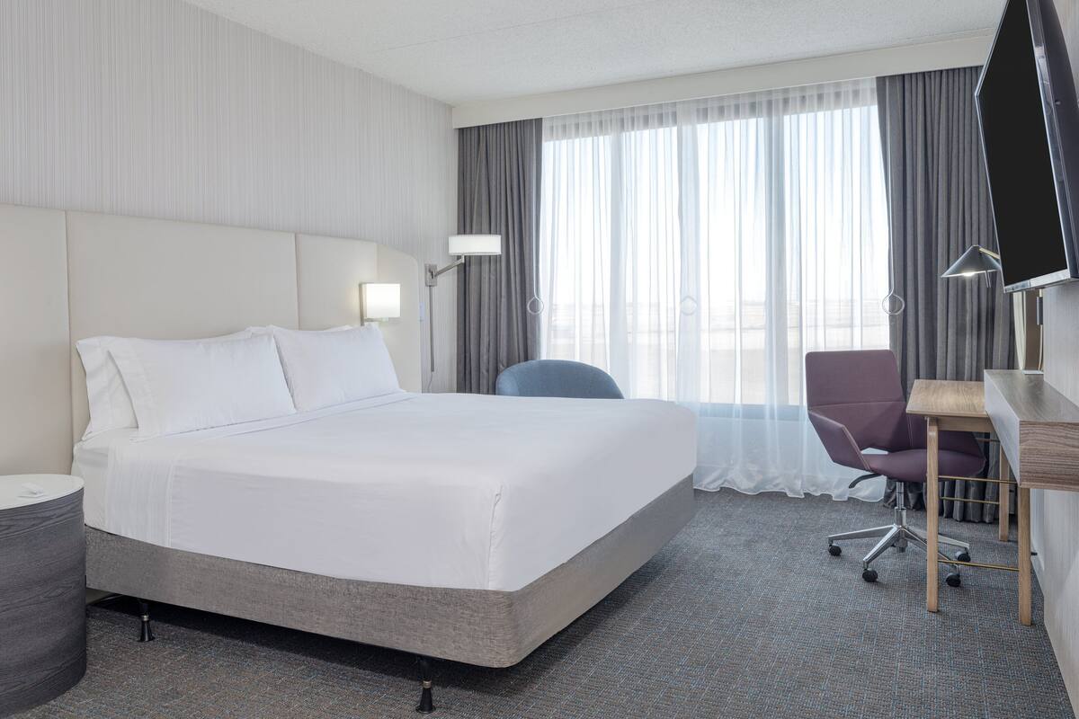 Ein Hotelzimmer mit einem großen Bett, einem Schreibtisch, einem Stuhl, einem Fernseher und einem Fenster mit Vorhängen.