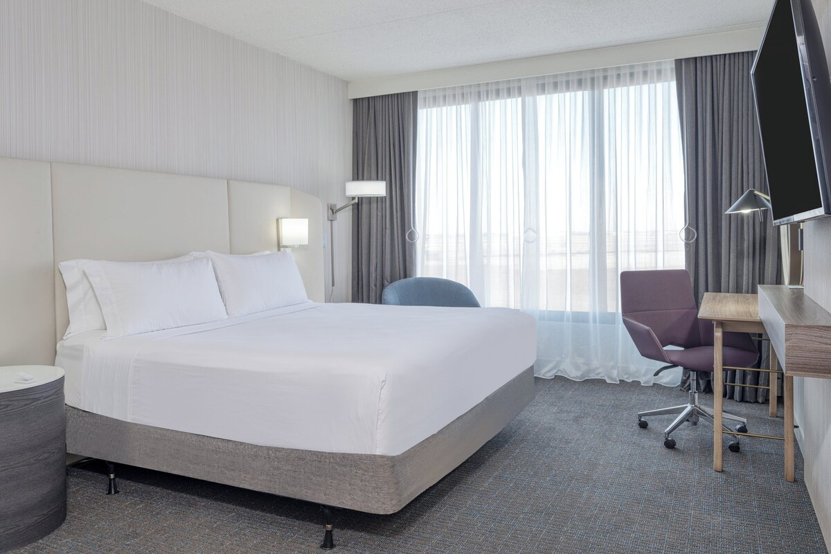 Une chambre d’hôtel avec un grand lit, un bureau, une chaise, une télévision et une fenêtre avec des rideaux.