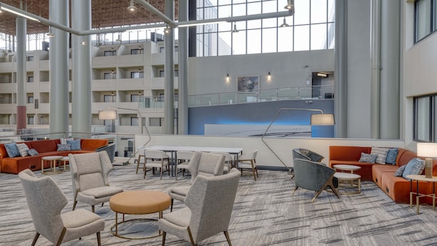 A spacious lobby with high ceilings, a central seating area with round tables and chairs, and a view of the building's exterior through large windows.