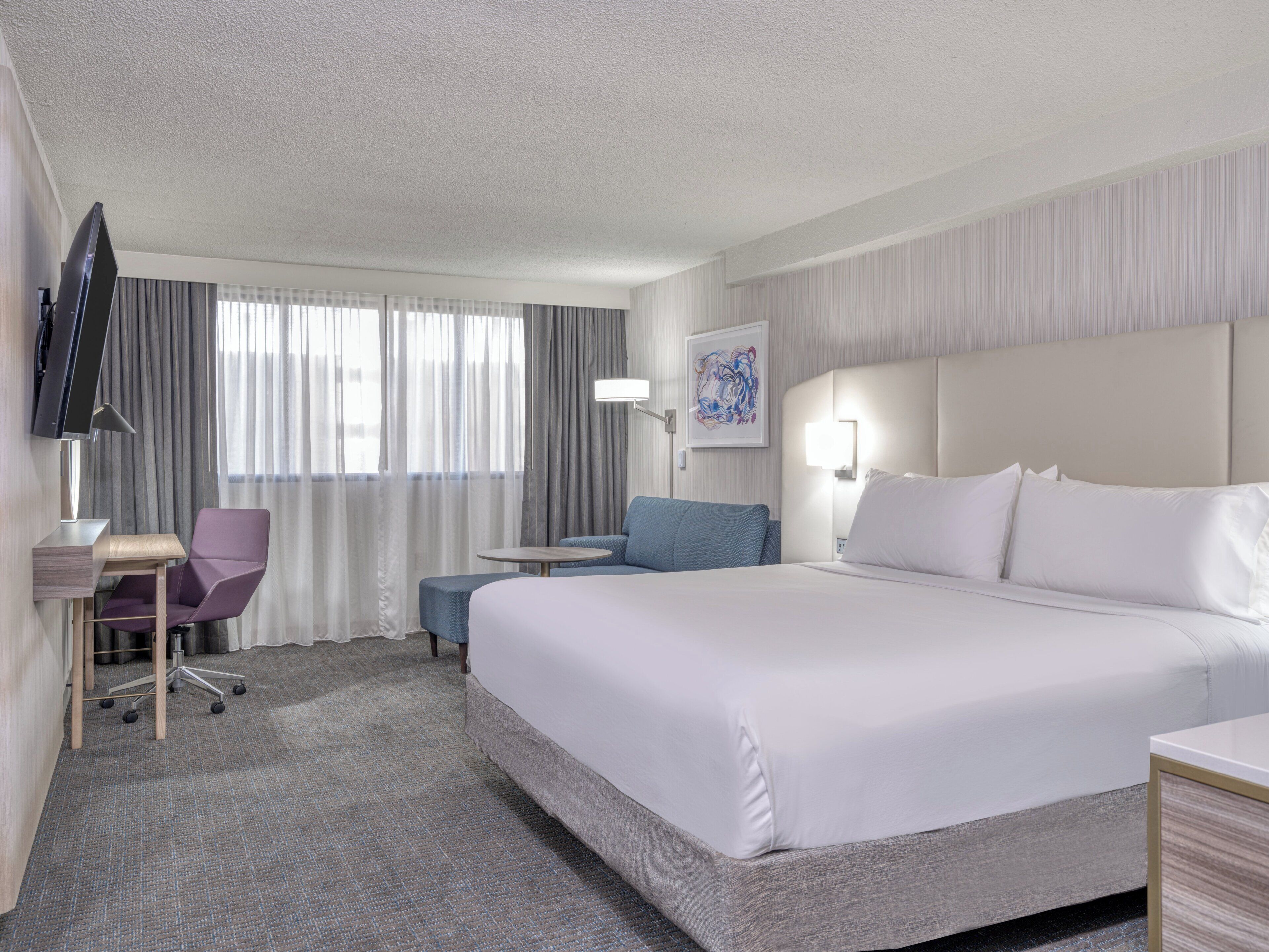A hotel room with a large bed, a desk with a chair, a flat-screen TV, and a window with curtains.