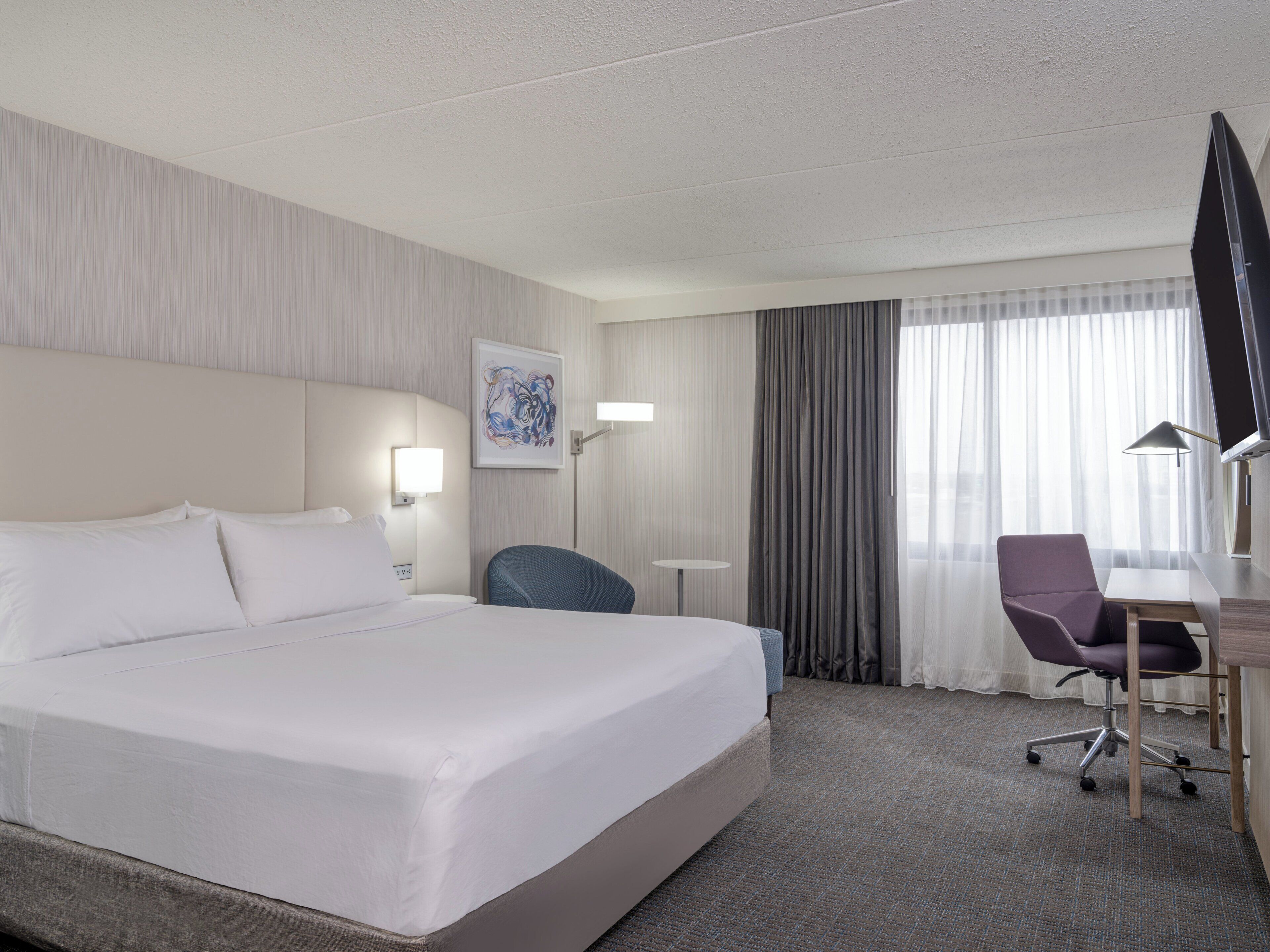 Una habitación de hotel con una cama grande, un escritorio con silla, un televisor de pantalla plana y un ventanal con cortinas.