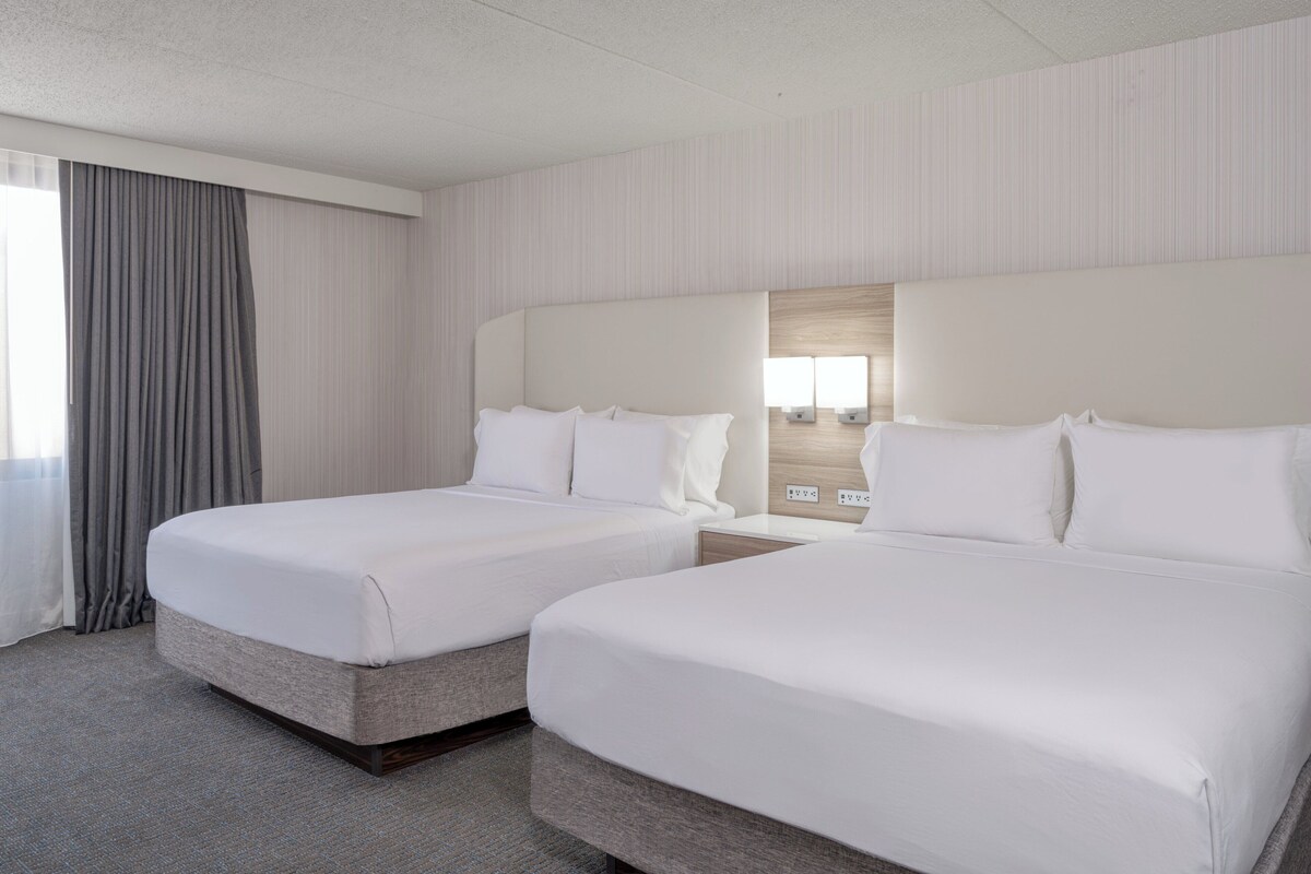 Une chambre d’hôtel avec deux lits, une grande fenêtre avec des rideaux et un luminaire fixé au mur.