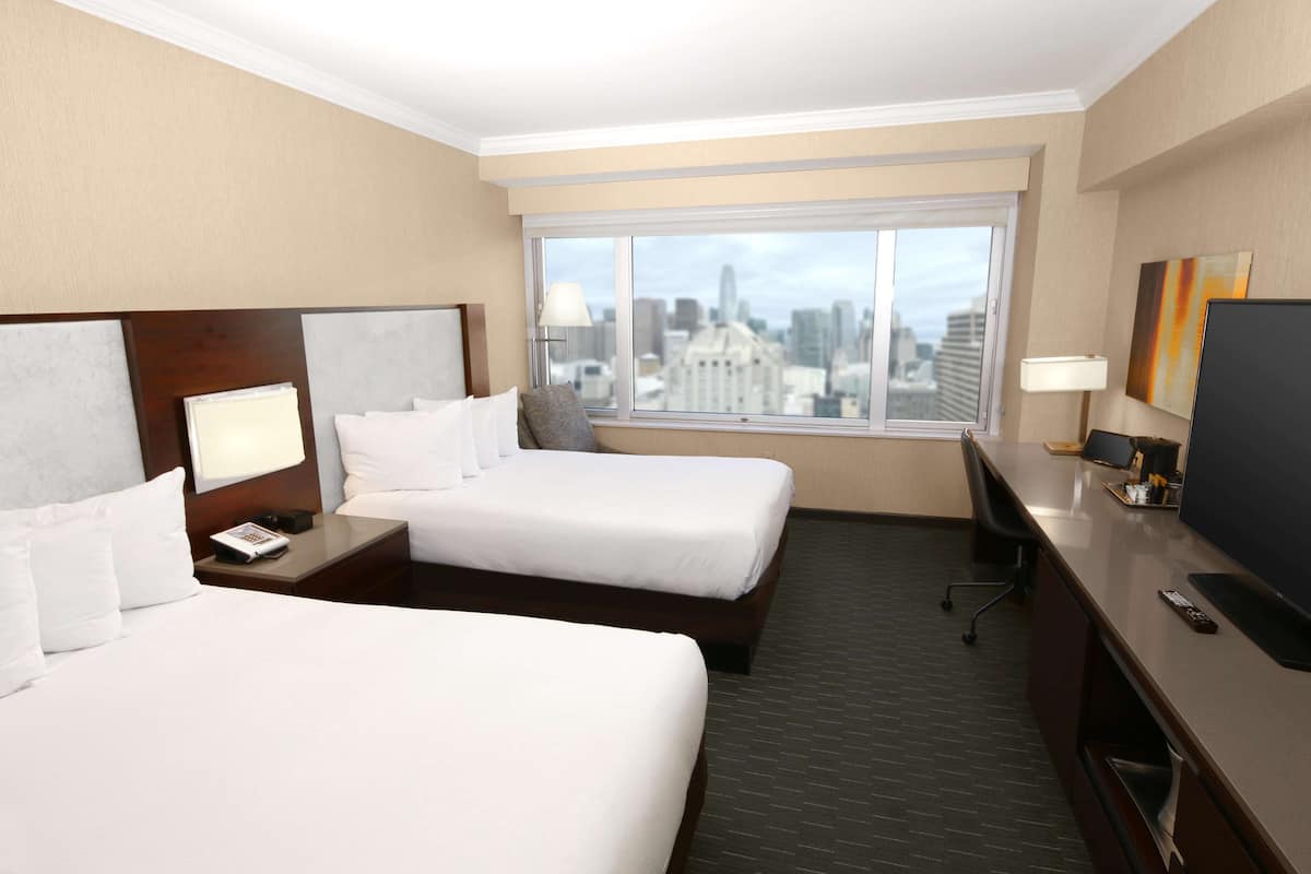 Habitación de hotel con dos camas, un escritorio, televisor y un amplio ventanal con vista a la ciudad.