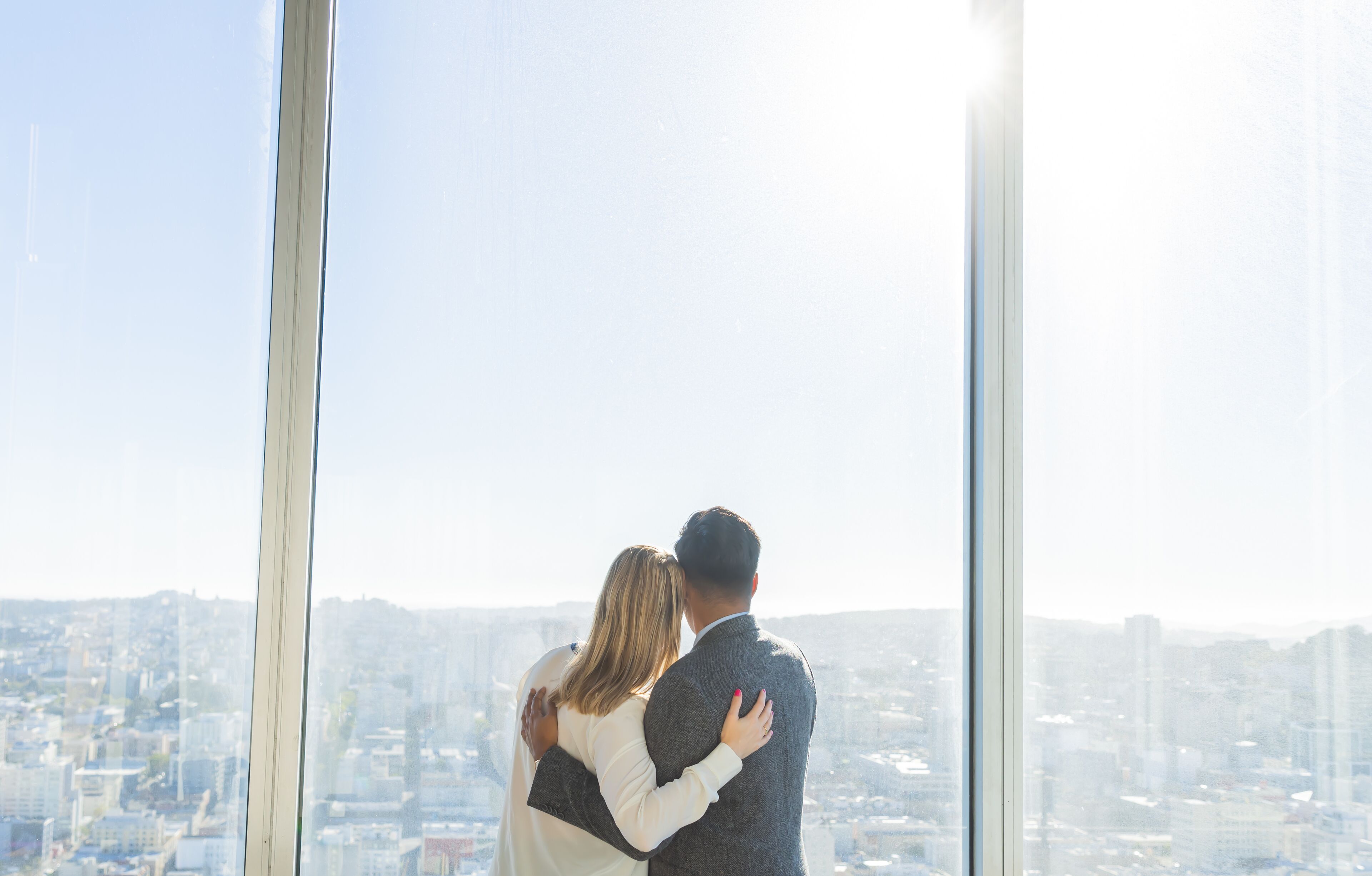A couple embracing in front of a large window overlooking a cityscape.