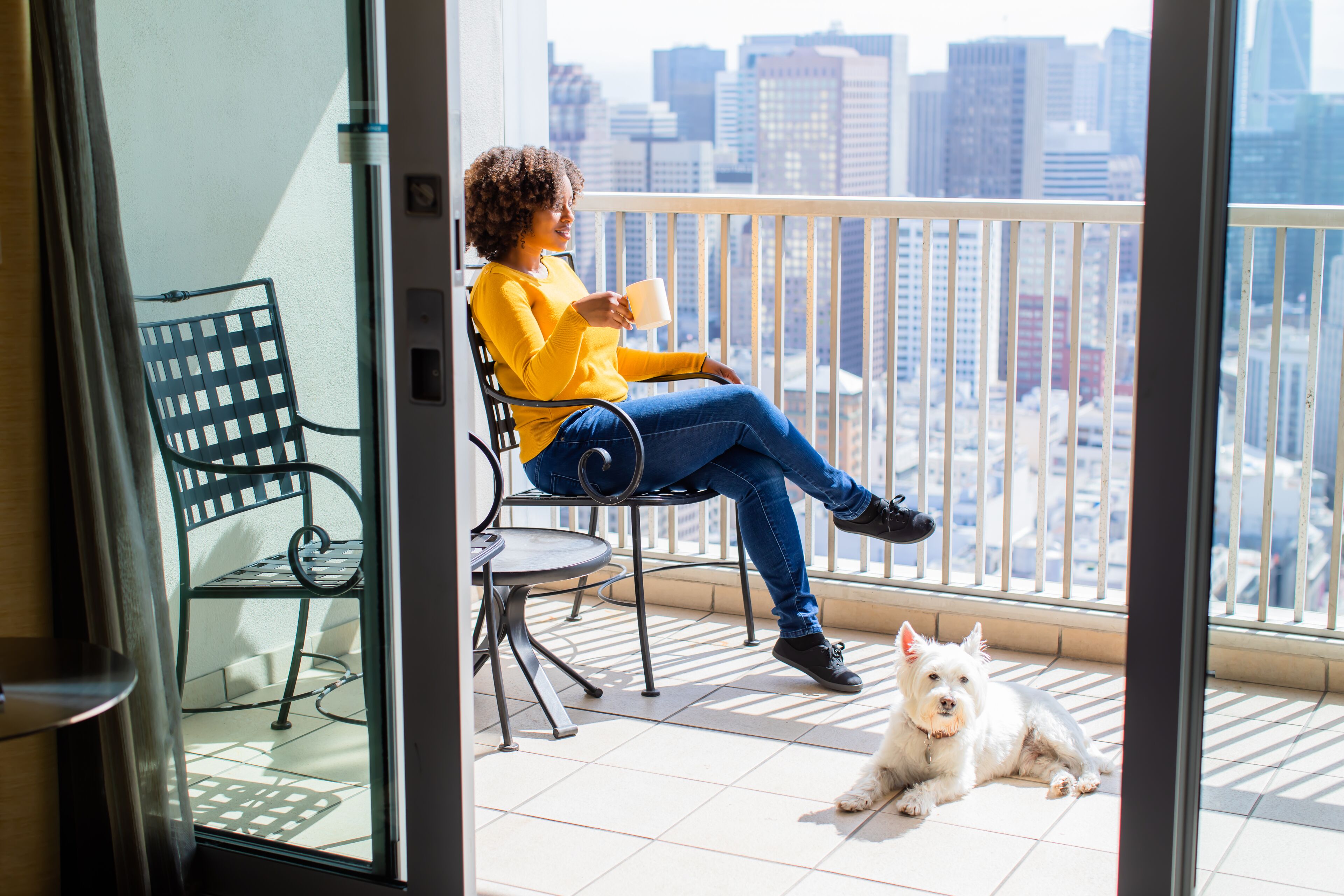 Una persona sentada en una silla de balcón con vista a la ciudad, una mesita pequeña y un perro en el suelo.
