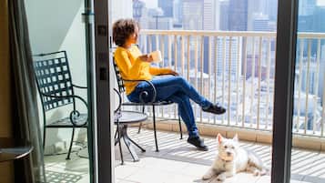 Una persona sentada en una silla de balcón con vista a la ciudad, una mesita pequeña y un perro en el suelo.