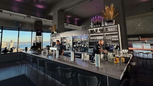 Un bar moderno con una barra larga, taburetes y un cantinero. Hay estantes con botellas y vasos, y una vista de la ciudad a través de amplias ventanas.
