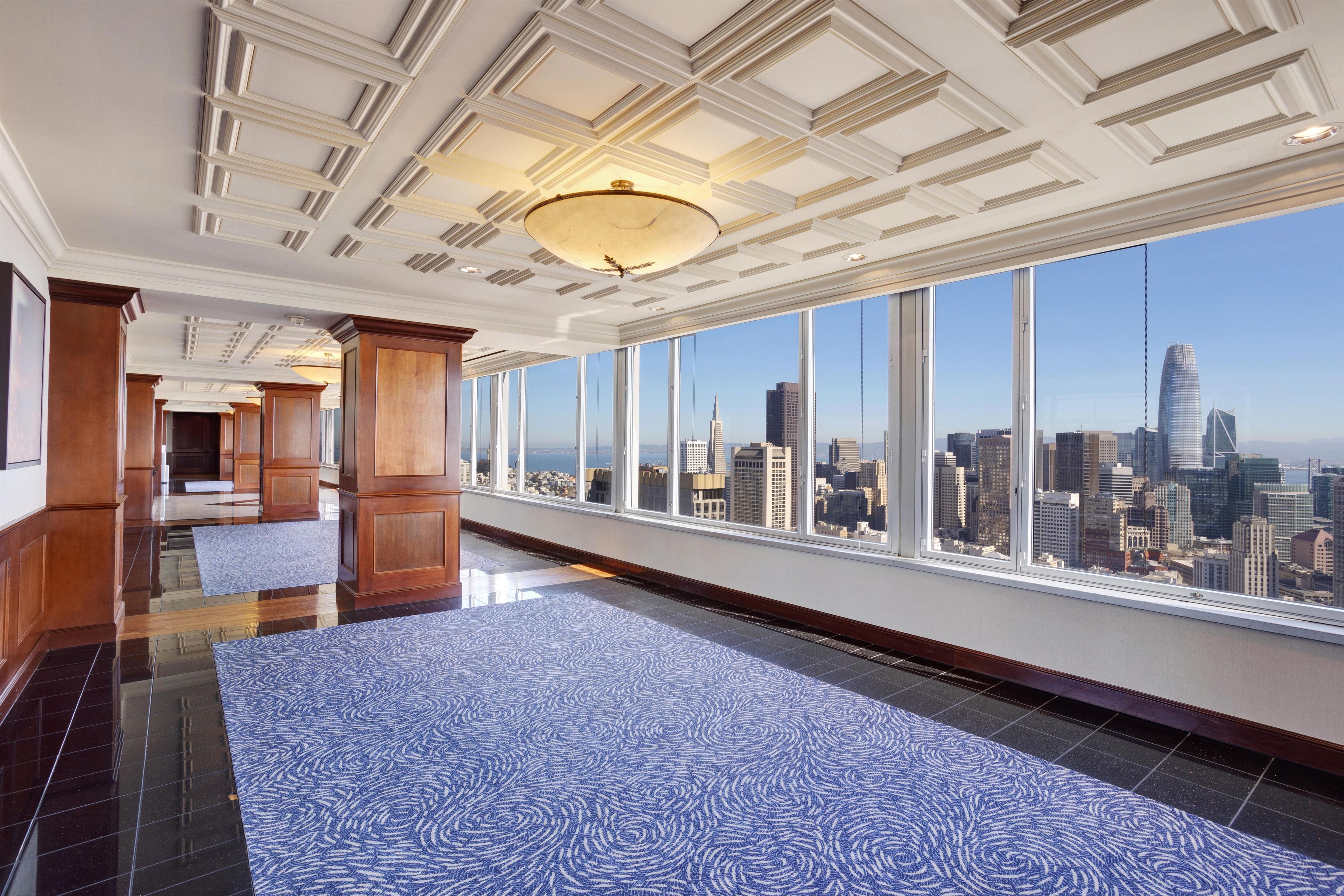 A spacious hotel corridor with a city view, wooden paneling, and a patterned carpet.