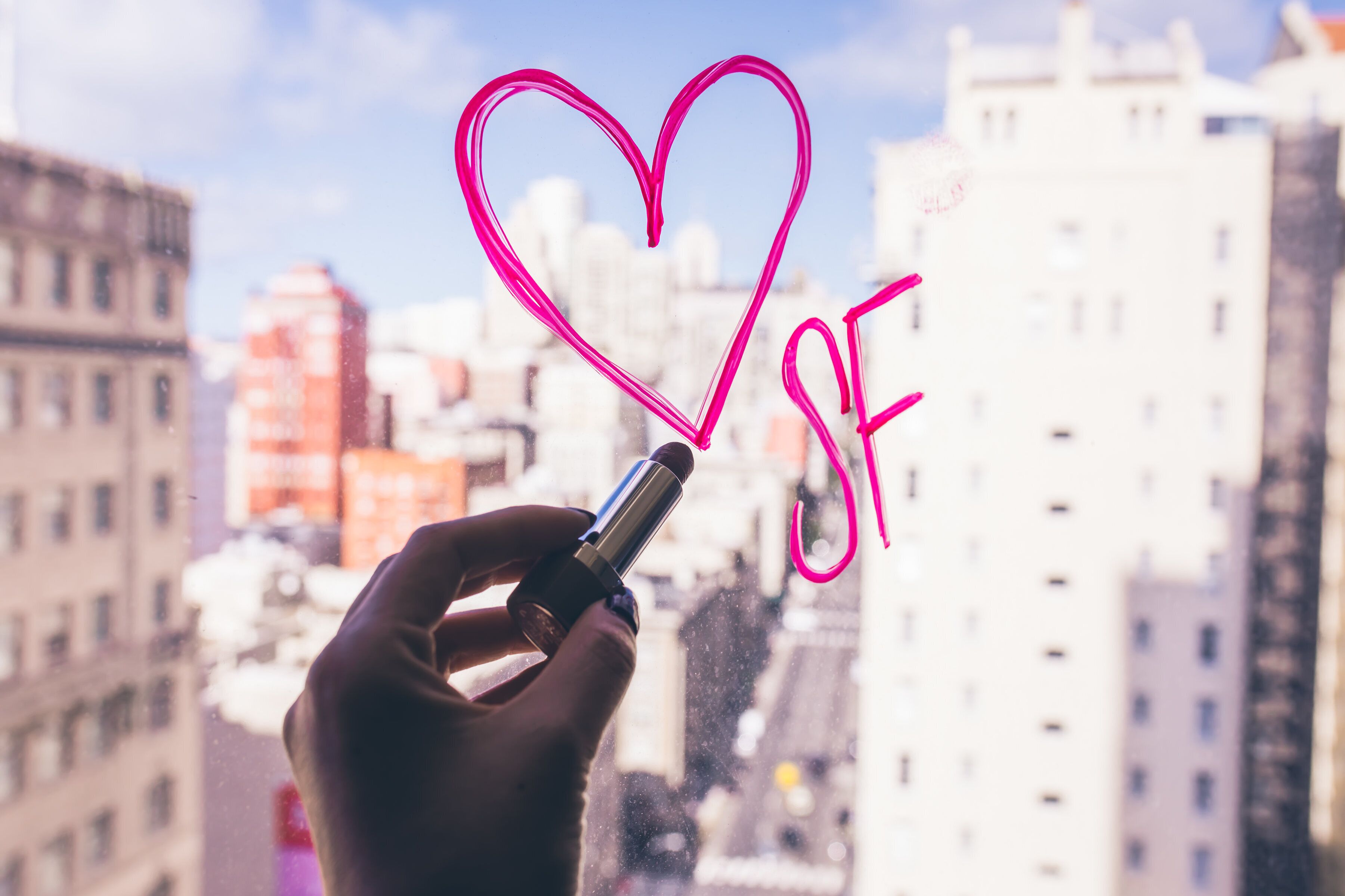 A hand holding a lipstick with a heart drawn on it, with a cityscape in the background.