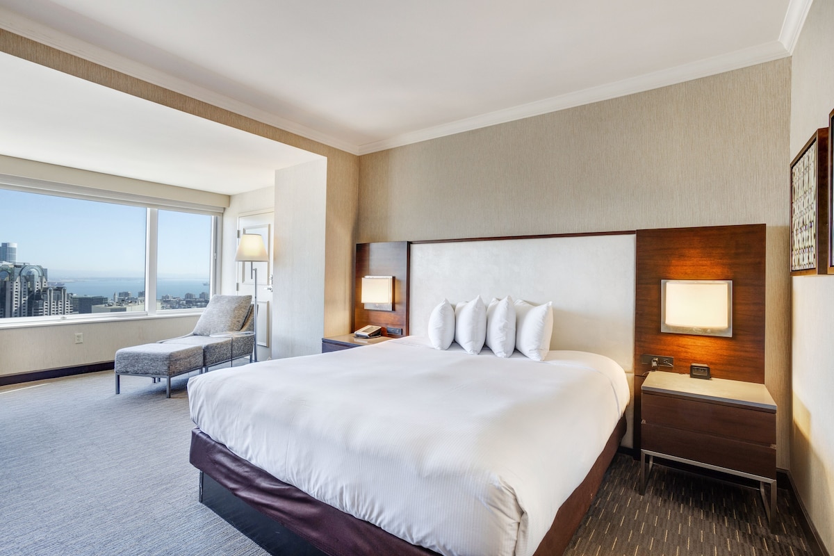 A hotel room with a large bed, a chair, a nightstand, and a view of the city.