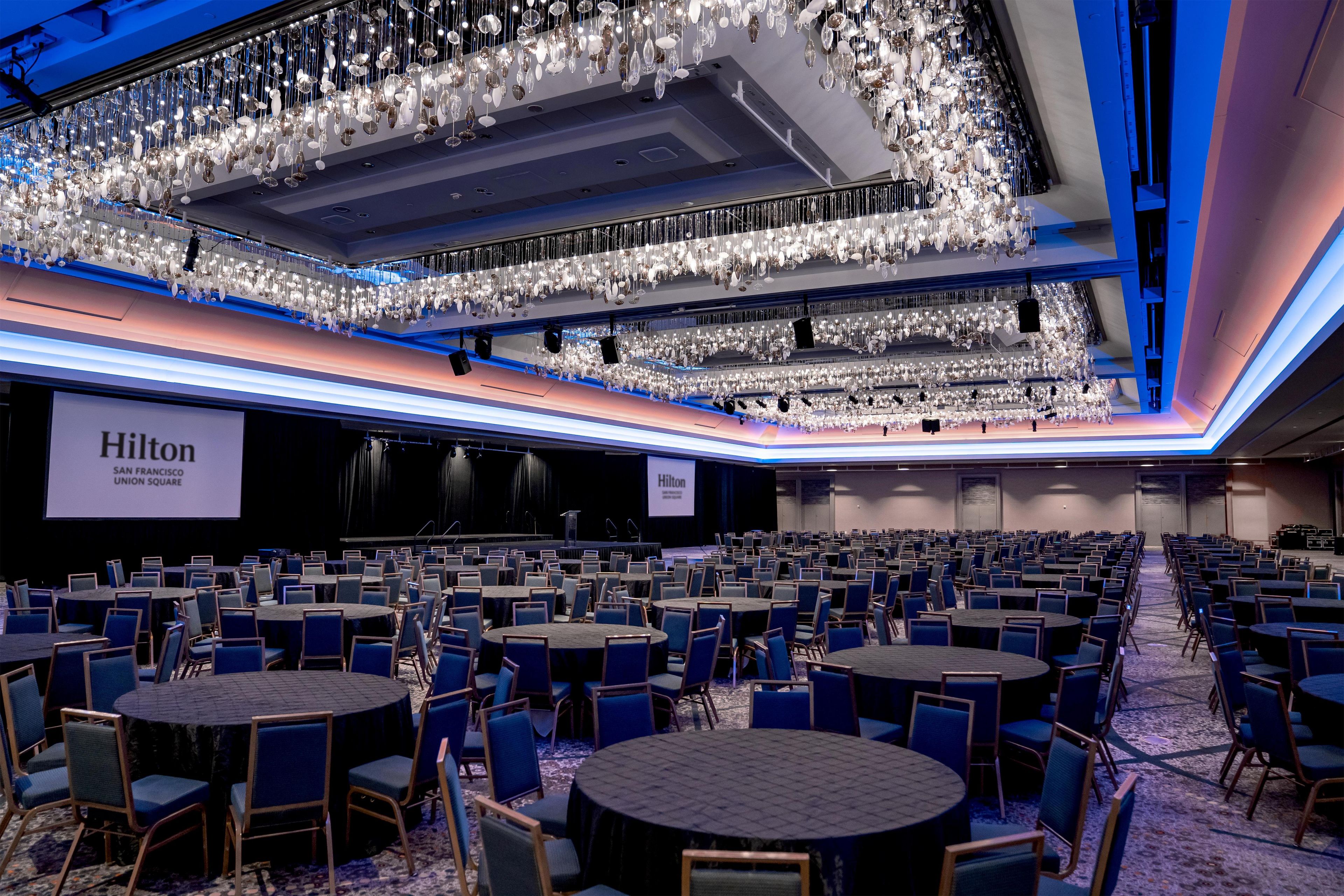Un amplio salón de conferencias con varias mesas y sillas, un escenario con un atril y lámparas de cristal colgantes en el techo.