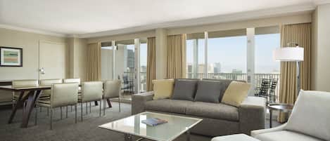 A hotel room with a sofa, armchair, dining table, and chairs. There is a large window with a city view.