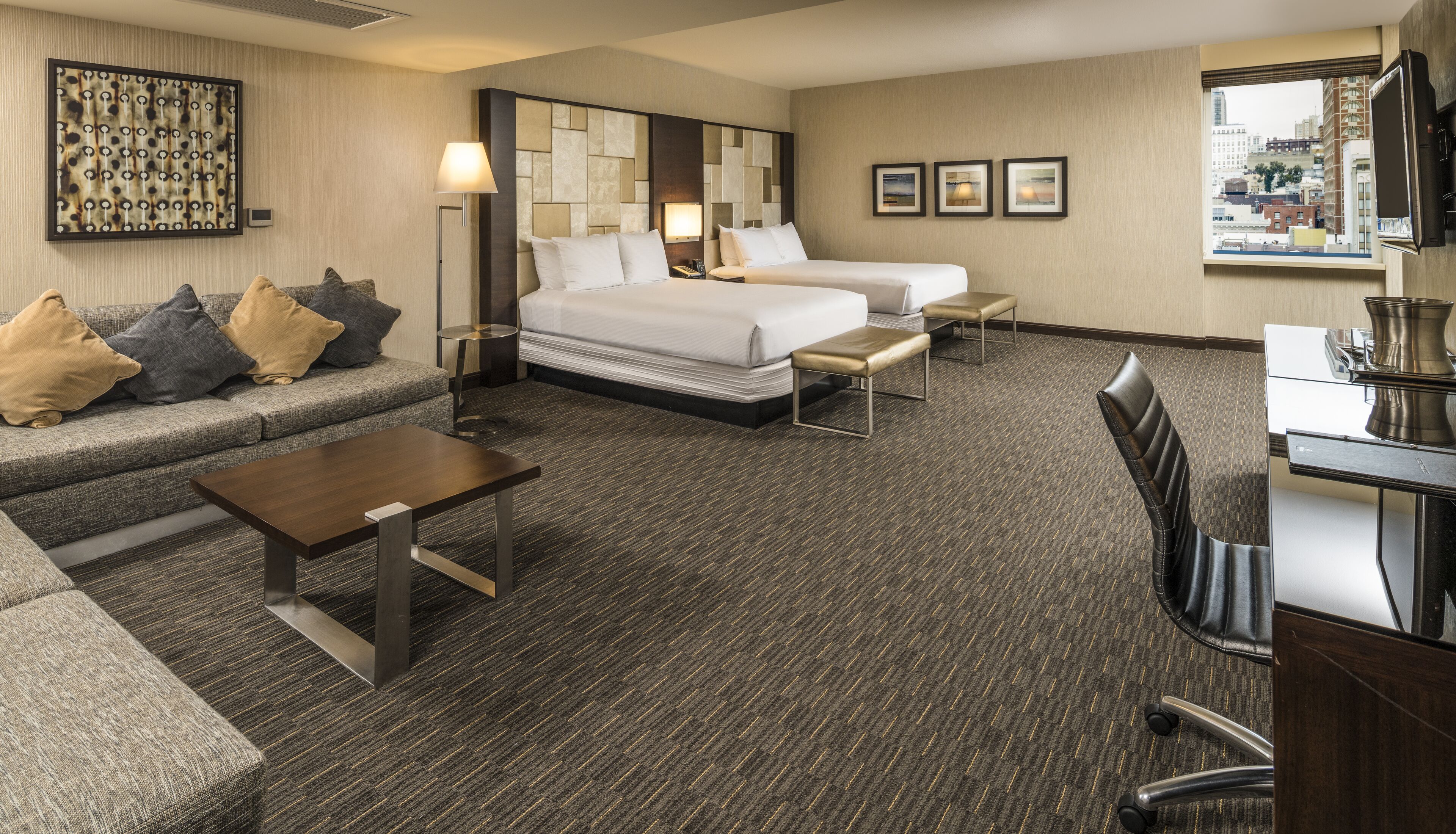 Habitación de hotel con una cama grande, un sofá, un escritorio y una silla. Hay tres cuadros en la pared y una ventana con vistas a la ciudad.