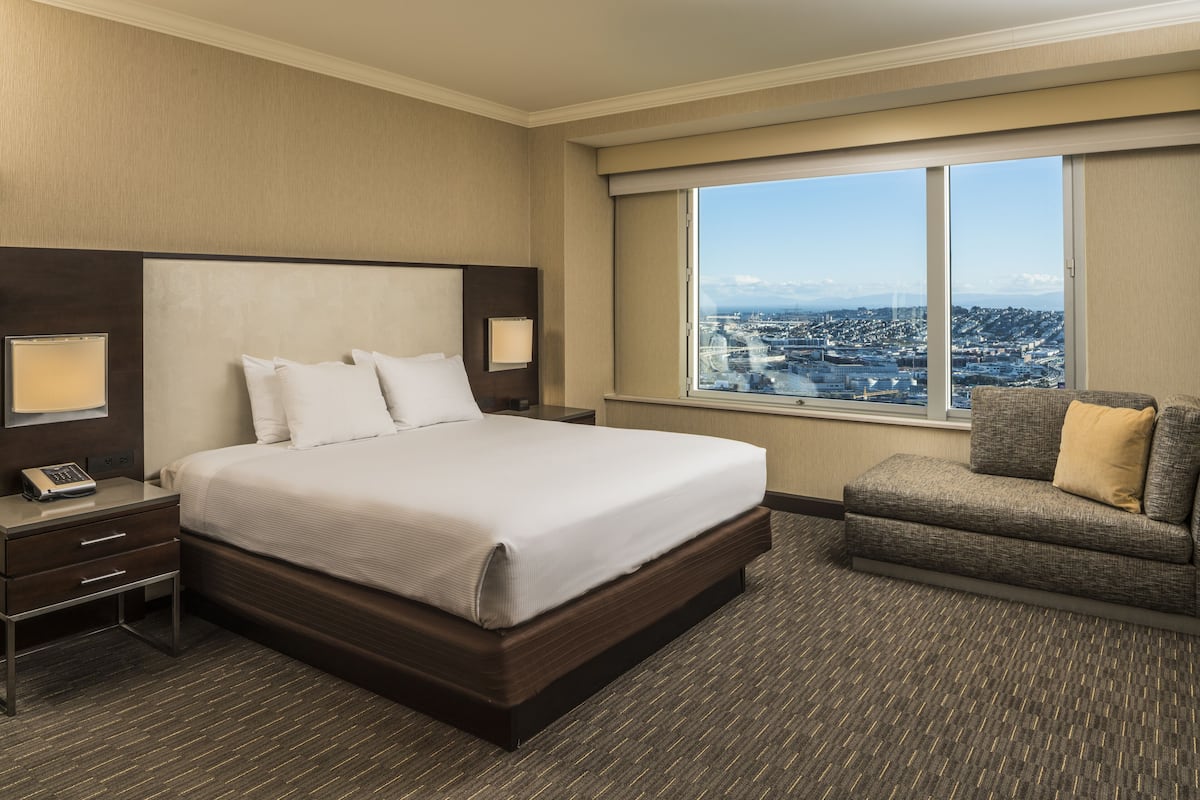 Habitación de hotel con una cama grande, una silla, una mesita de noche y un ventanal con vistas a la ciudad.