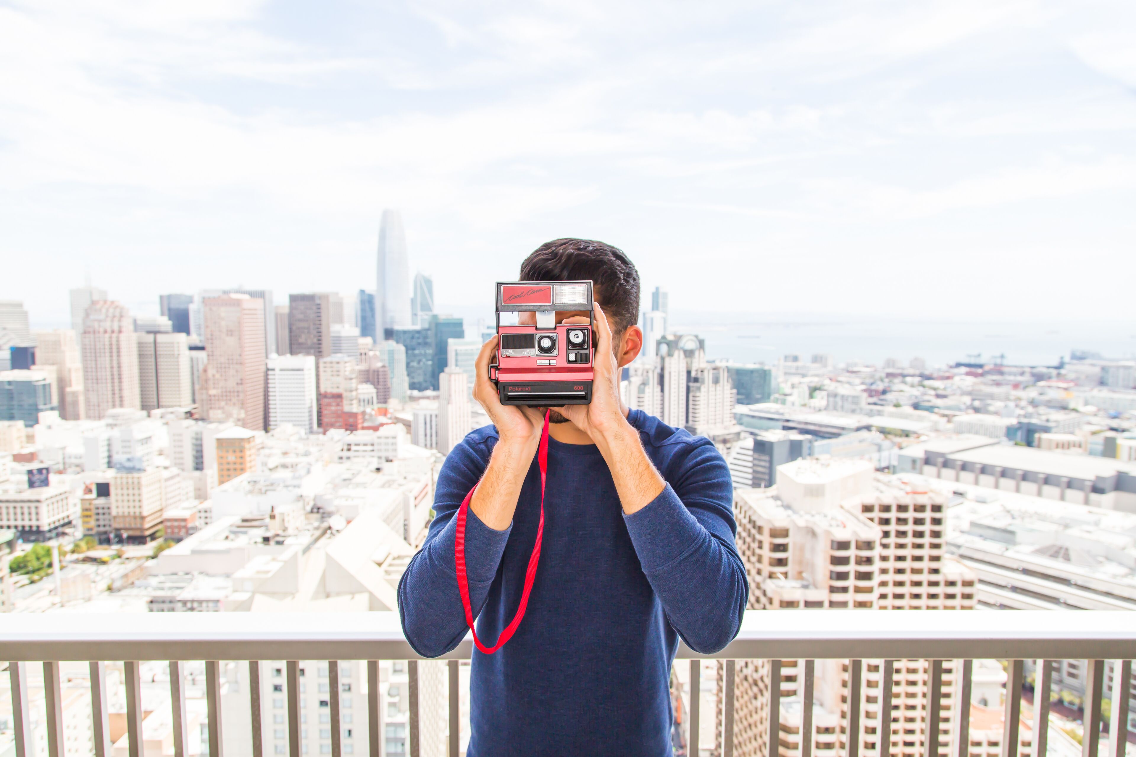A person holding a vintage camera with a red strap, overlooking a cityscape with numerous buildings and a clear sky.
